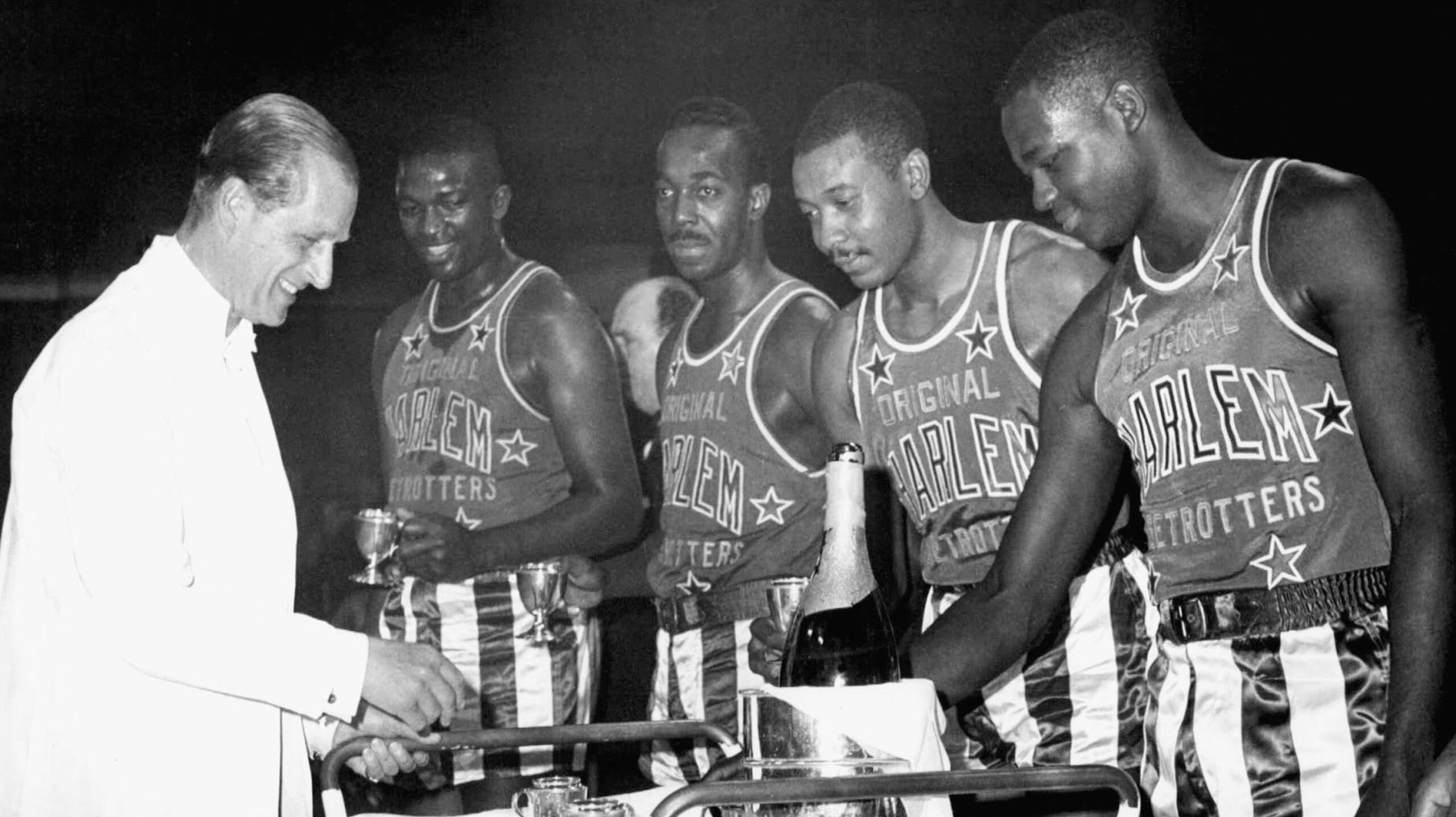 Members of the Harlem Globe Trotters Professional Basketball Team are served champagne by Prince Philip, the Duke of Edinburgh, on Nov. 4, 1963 at a London Hotel. Trotters flew to the British capital to play the "Lord Taverners" team in a charity game. Prince Philip is the 12th man on the British team. - Associated Press